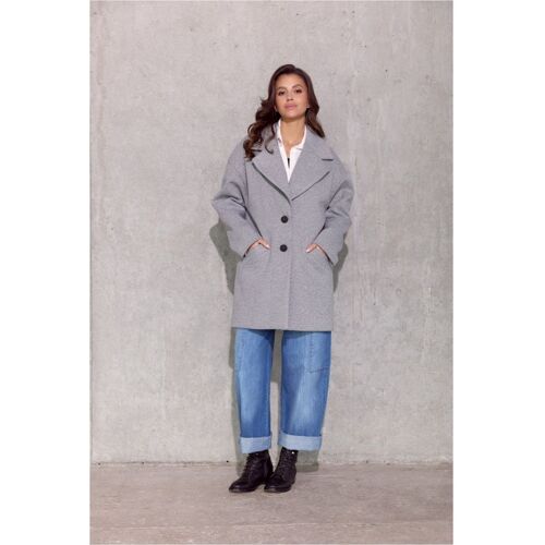 Frau trägt grauen Mantel, blaue Jeans und schwarze Stiefel vor grauer Wand.