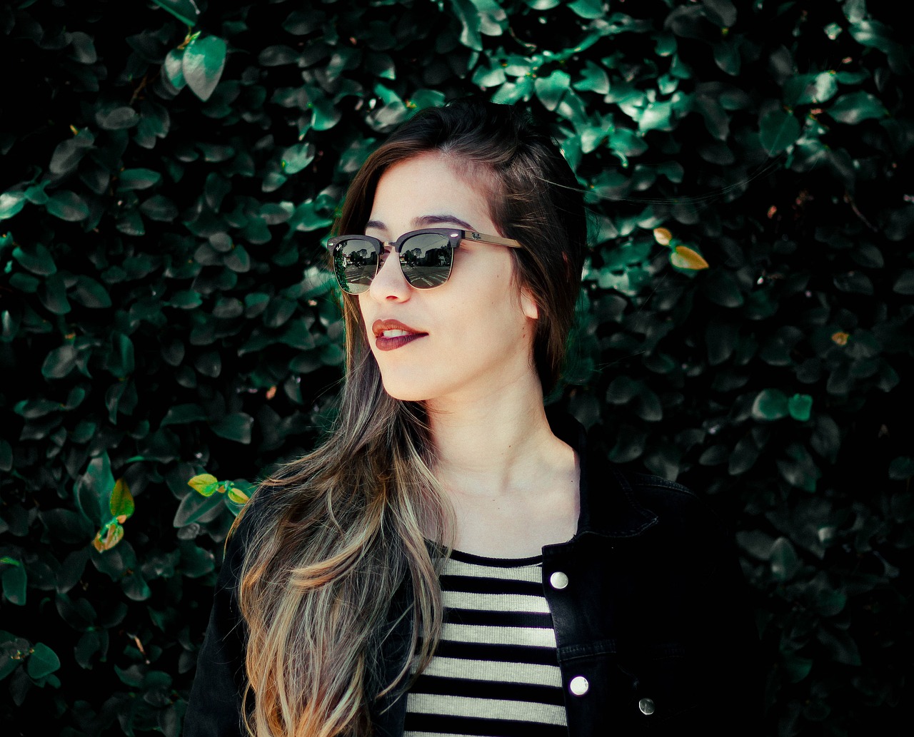 Frau mit Sonnenbrille steht vor einer grünen, blätterbedeckten Wand.