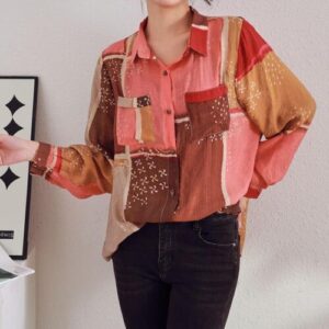 Person trägt ein buntes Patchwork-Hemd und schwarze Jeans vor einer weißen Wand.