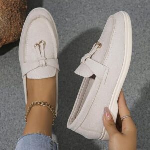 Beige Loafers mit Schnalle auf grauem Boden und einer Hand im Bild.