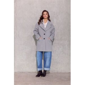 Frau trägt grauen Mantel, blaue Jeans und schwarze Stiefel vor grauer Wand.