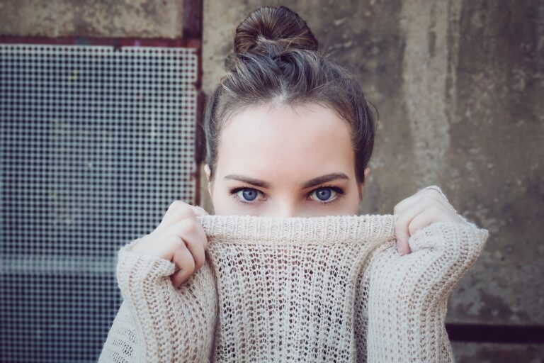 Person verdeckt Gesicht teilweise mit hellem Strickpullover vor metallischer Gitterwand.